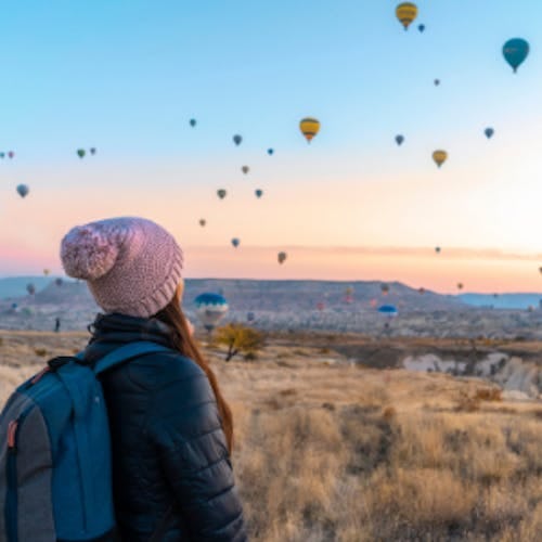 A woman staring up at hot air balloons at sunset over a desert landscape 