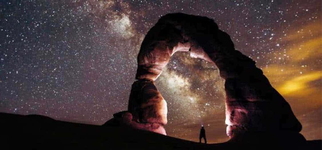 A small silhouette of a stargazer looking up through a stone arch in the Arches National Park, with a brilliant view of the stars above