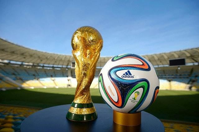 A golden Fifa World Cup trophy next to the white football on a stand with the stadium in the background 