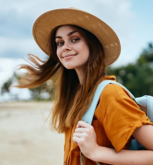 A happy woman with a backpack and sunhat walking along a beach, looking back at the camera and smiling 