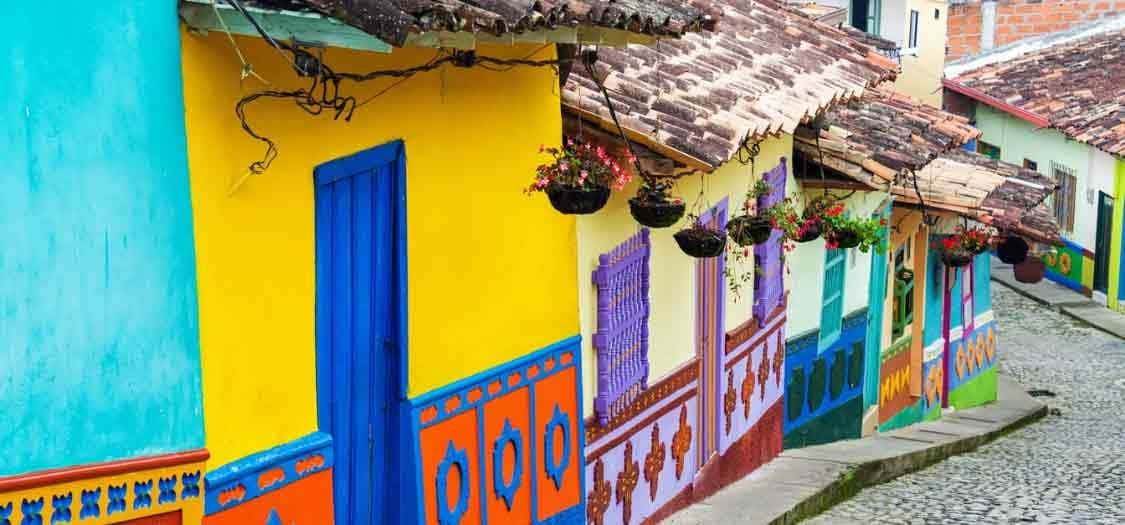 A street view depicting the colours of traditional homes in Bogata, Colombia, in vivid bright yellows, reds, greens and blues. The short buildings have red tiles and hanging plant pots