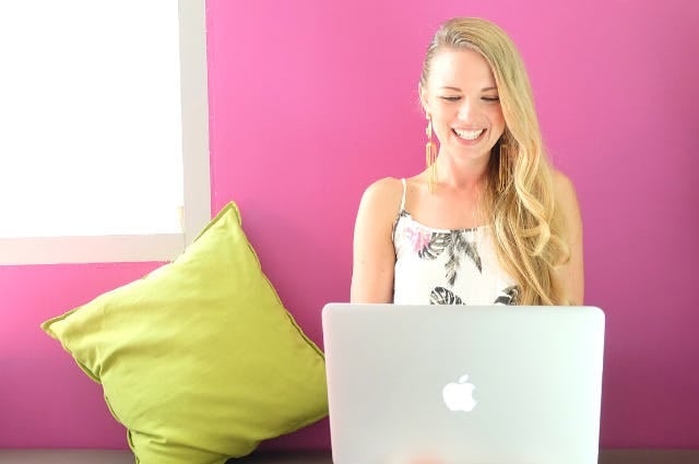 Blonde woman in a sundress working on her laptop against a pink wall