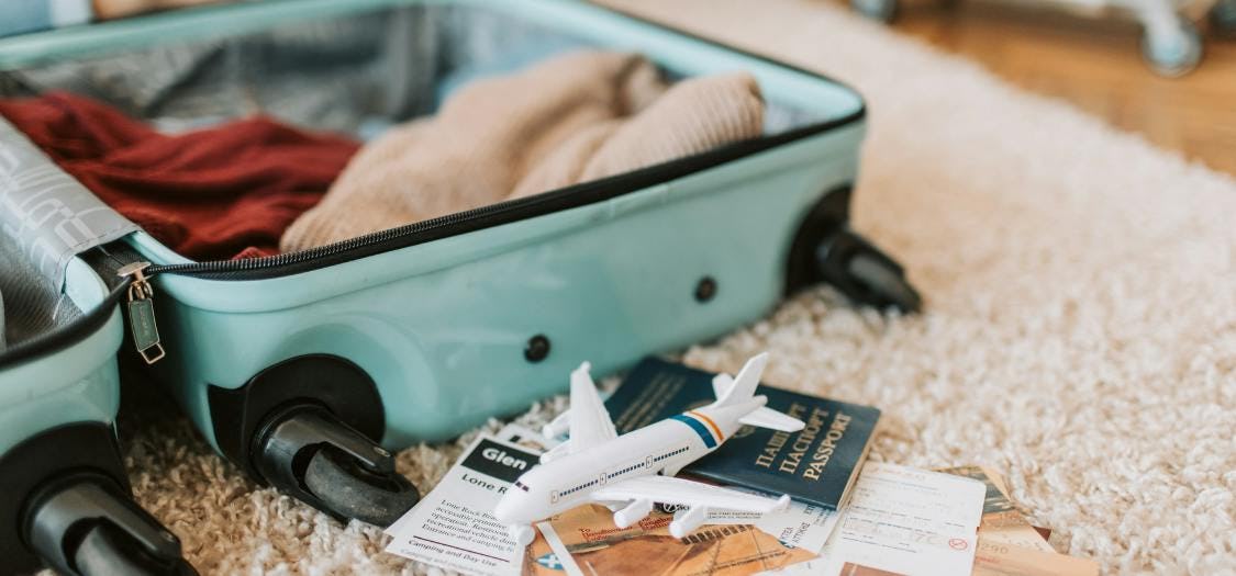 A low shot of an open teal suitcase on a shaggy cream rug. There's items starting to be packed, including a red jumper. Travel documents including a passport and small model of a plane are in front 