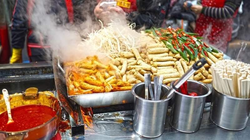 A shot of a street food vendor, with a large glass of hot red sauce, chillies and veg above a hot grill 