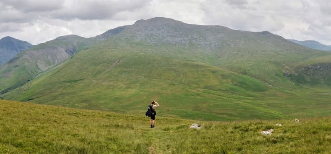 A wide shot of a man walking though a green landscape with rocky hills