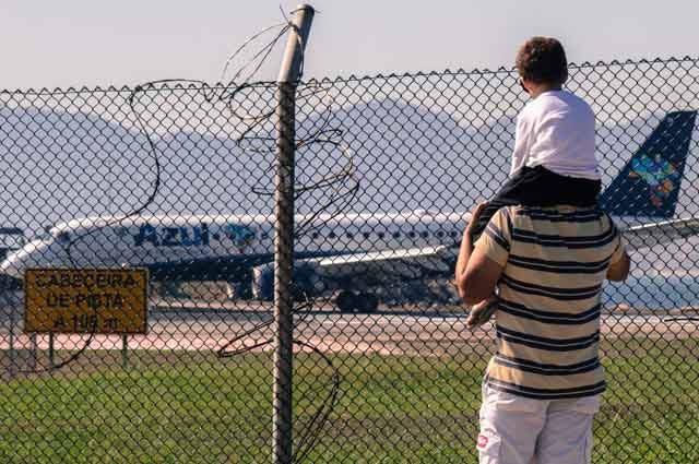 A man with a small child sat on his shoulders while they watch an aircraft on an airport runway 