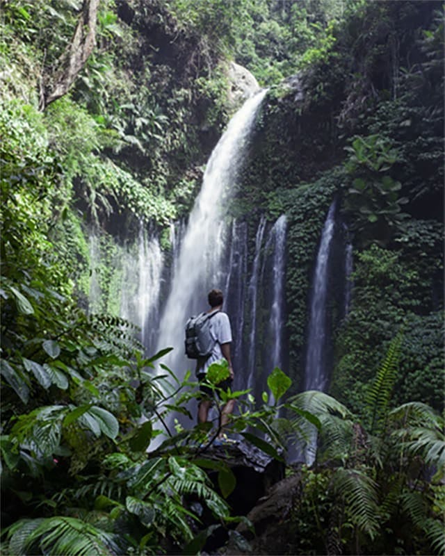 A luscious green environment with waterfalls and a backpacker in the foreground looking up at the falls