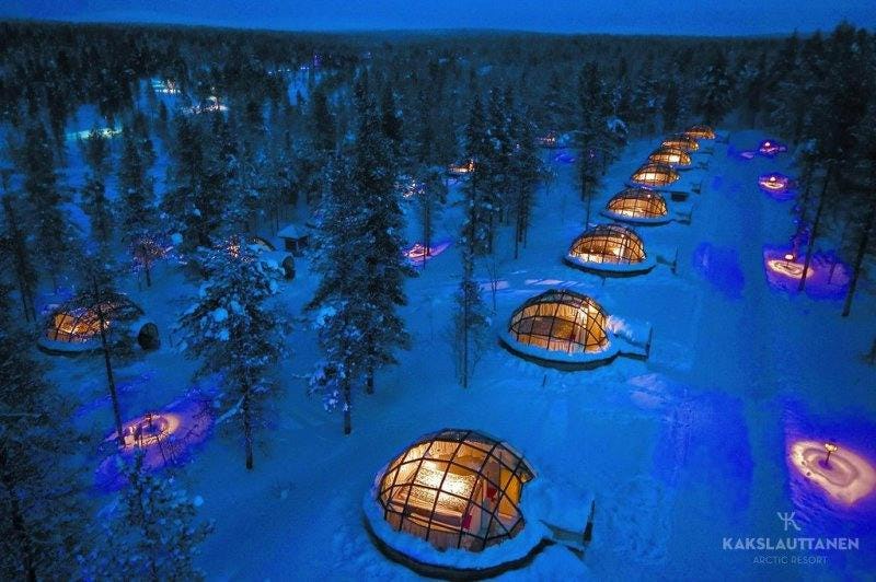 A shot taken in an arctic resort, taken looking down a glass dome structures that are available to rent. Each dome is lit by warm light in contrast to the cold snow and dark pine trees outside