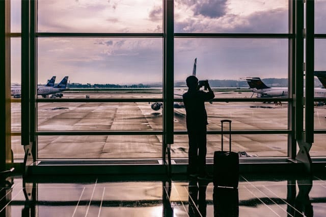 Silhouette of a person standing inside an airport taking a picture of planes on the runway through a window