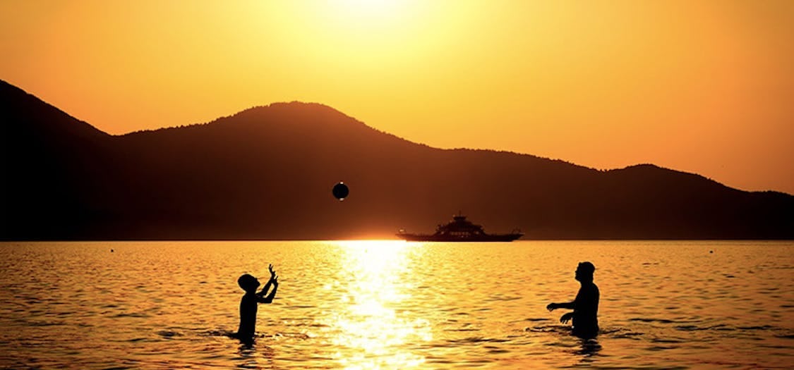 2 people playing with a beach ball in the sea, with mountains and a sunset in the distance