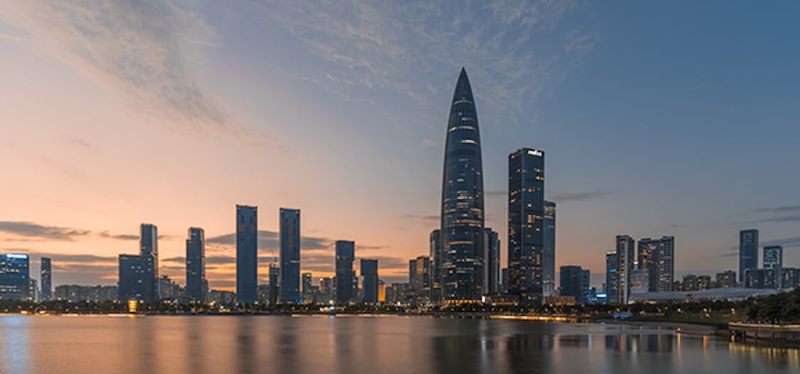 The futuristic skyline of Shenzhen bathed in dim glow from the setting sun casting reflections over the water below
