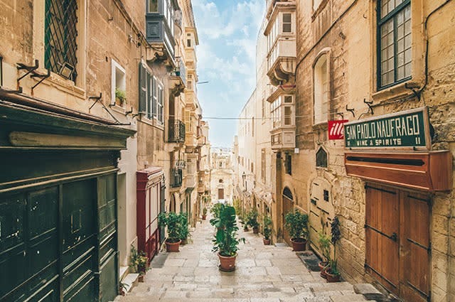 A narrow cobbled street in Malta, highlighting the old age of the area. Buildings feature wooden shutters and the the street is aligned with potplants
