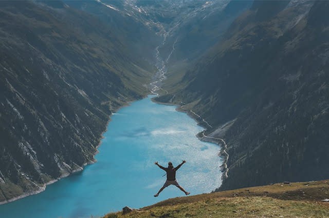 Man star jumping on a grass bank above a large canyon overlooking a lake