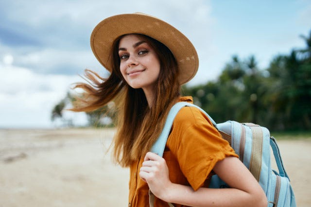shot of a woman in a sunhat walking along a beach while turning back to look at the camera with a smile on her face