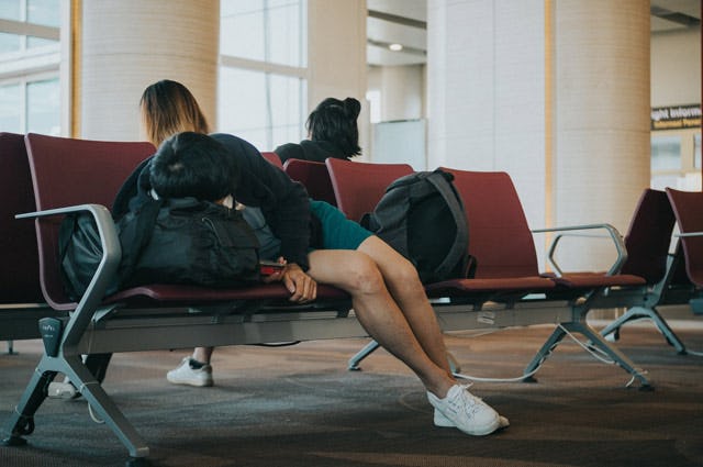Picture of a person sleeping at an airport on a bench of chairs, using their bag as a pillow